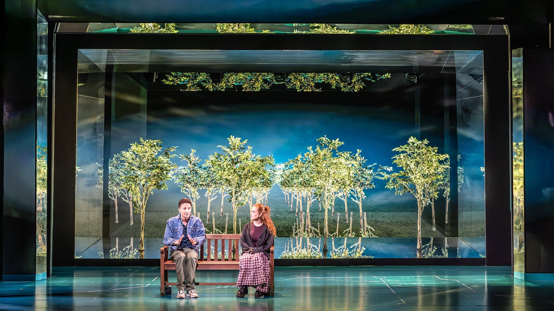 Two people sitting on stage on a bench in from of a forest background 